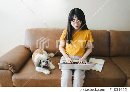 Work from home concept. Happy young asian woman in glasses using laptop while sitting on couch with her cute dog. 74831635