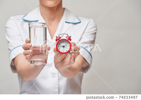 Healthy eating or lifestyle concept - female woman doctor holding and a glass of clear fresh waterand small red alarm clock Healthy eating or lifestyle concept - female woman doctor holding and a glass of clear fresh waterand small red alarm clock 74833497