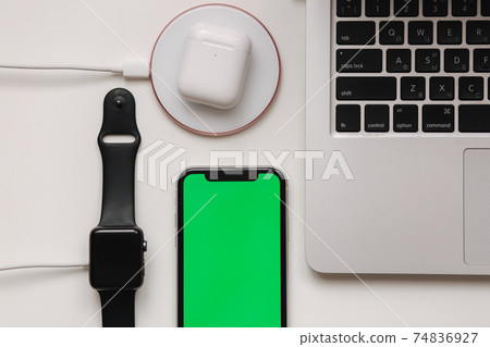 Charging of different devices on working place. Few cabel for wireless charging. Laptop, smart watch, smart phone with green screen and witeless headphones on white table. Top view. Mock up. Place for Charging of different devices on working place. Few cabel for wireless charging. Laptop, smart watch, smart phone with green screen and witeless headphones on white table. Top view. Mock up. Place for 74836927