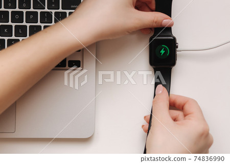 Female hand put smart watch on wireless charging with on-screen charging indicator. Work place near at the laptop. Top view Female hand put smart watch on wireless charging with on-screen charging indicator. Work place near at the laptop. Top view 74836990
