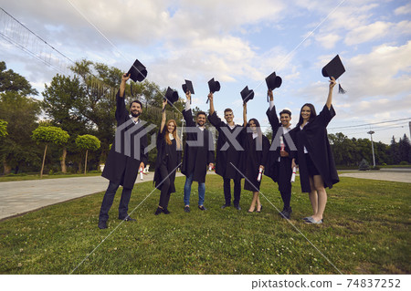 University graduate students raising mortarboards up outdoors. College grads in gowns celebrating commencement day 74837252