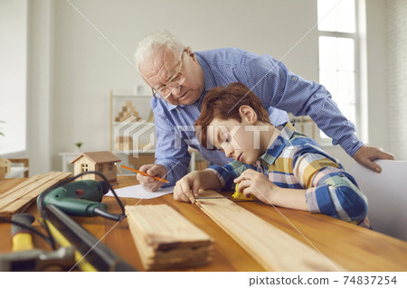 Grandfather helping his grandson to measure wooden plank at the carpentry workshop Grandfather helping his grandson to measure wooden plank at the carpentry workshop 74837254