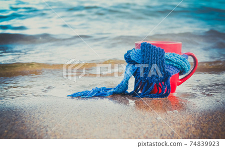 Red mug with coffee tied with blue knitted scarf stands on sandy beach 74839923
