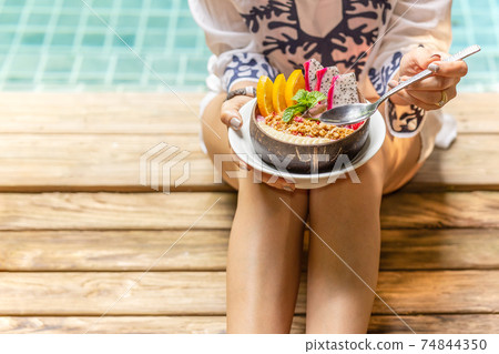 Woman holding smoothie bowl with mixed fruits and granola by swimming pool. Woman holding smoothie bowl with mixed fruits and granola by swimming pool. 74844350