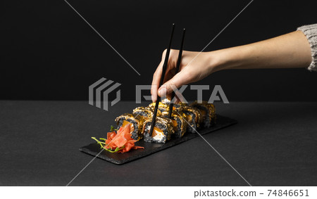 Hand with black chopsticks wants to take custom sushi roll with cuttlefish ink, salmon, cream cheese, pepper pumpkin, eel and edible gold leaf with ginger, wasabi on a square plate on black table. 74846651