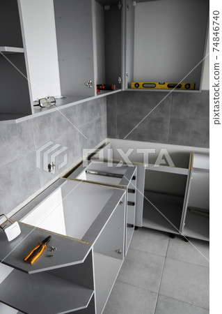 Custom kitchen cabinets installation without a furniture facades mdf. Gray modular kitchen from chipboard material on a various stages of installation in kitchen with a grey tile on floor and walls. 74846740