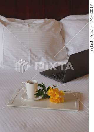 Cup of tea with a yellow flower on a white bed next to laptop. Cup of tea with a yellow flower on a white bed next to laptop. 74850017