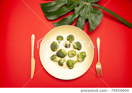 Plate of fresh raw broccoli with cutlery placed on red table near Monstera leaf 74850604