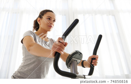 Attractive fit woman exercising on a spin bike at home on a beautiful sunny day. Cardio workout. Fitness at home. Closeup Attractive fit woman exercising on a spin bike at home on a beautiful sunny day. Cardio workout. Fitness at home. Closeup 74850816