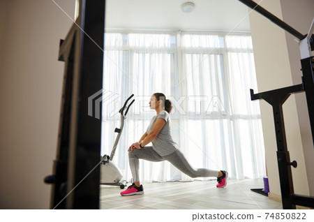 Side full-length portrait of young sporty woman in gray sportswear performing lunges against large windows at home 74850822