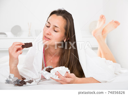 Adult woman secretly eating chocolate under sheets in bed 74856826