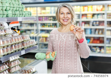 Mature woman choosing fresh eggs in grocery shop Mature woman choosing fresh eggs in grocery shop 74856974