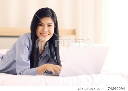 Young and beautiful Asian woman laying on a bed in bedroom with friendly smile and looking to camera and using notebook laptop computer with happiness manner. Concept for modern woman lifestyle 74860094