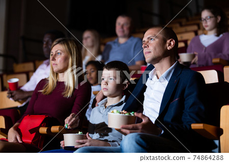 Teenage son watches movie with his parents and eats popcorn in cinema hall Teenage son watches movie with his parents and eats popcorn in cinema hall 74864288