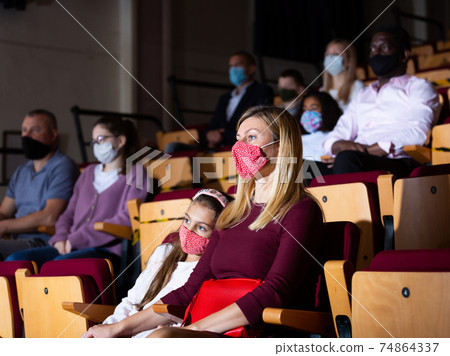Mom and daughter in protective masks in auditorium watching play in theater Mom and daughter in protective masks in auditorium watching play in theater 74864337