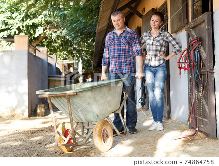 Smiling couple with barrow standing at horse stable outdoor Smiling couple with barrow standing at horse stable outdoor 74864758