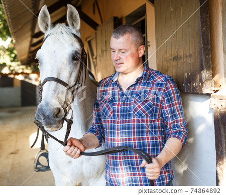 Positive male farmer standing with white horse at stable Positive male farmer standing with white horse at stable 74864892