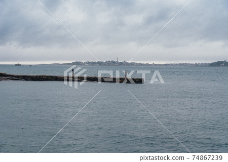 Back view of two adults standing on pier facing to the calm sea against the Island of Batz 74867239