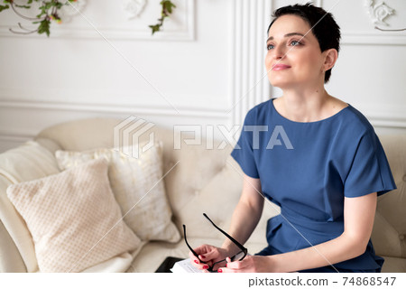 Pretty young brunette woman sits on sofa holding eyeglasses in her hand looking up 74868547
