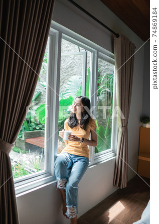 Attractive asian girl holding a cup with hot tea or coffee and looking at the window in her home and having a perfect cozy morning. Attractive asian girl holding a cup with hot tea or coffee and looking at the window in her home and having a perfect cozy morning. 74869184