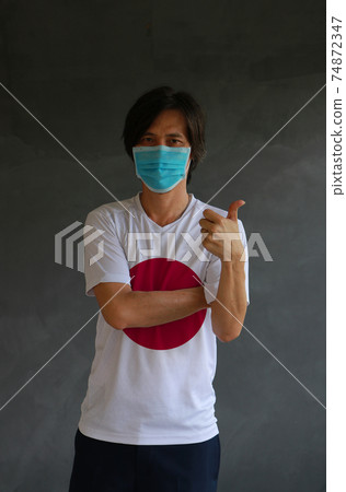 Man wearing hygienic mask and wearing Jananese flag colored shirt and cross one s arm with thumbs up Man wearing hygienic mask and wearing Jananese flag colored shirt and cross one s arm with thumbs up 74872347