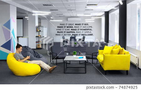 Creative space.With young man with smartphone sitting in yellow bean bag in the first plan and Conference , meeting table and office armchair on the second plan. With motivation words on the wall 74874925