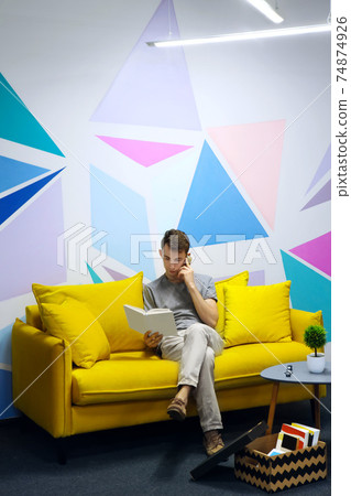 Young man sitting on comfortable yellow sofa and speak on telephone, hold book in hand. Against colored wall with geometrical figure.Coffee table with green plant near sofa. Relaxing zone.Copy space Young man sitting on comfortable yellow sofa and speak on telephone, hold book in hand. Against colored wall with geometrical figure.Coffee table with green plant near sofa. Relaxing zone.Copy space 74874926