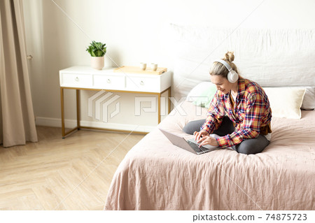 Working from home. Ordinary caucasian woman in casual wear and headset sits on the bed and using laptop, selective focus. 74875723
