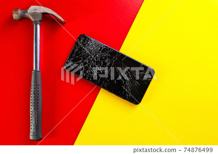 A broken black smartphone with a cracked display and a metal hammer lies on a red and yellow A broken black smartphone with a cracked display and a metal hammer lies on a red and yellow 74876499