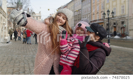 Women tourists taking selfie photos on mobile phone with adoption child girl on winter city street Women tourists taking selfie photos on mobile phone with adoption child girl on winter city street 74878025