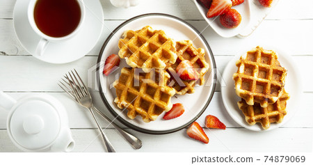 Delicious breakfast. Belgian waffles with honey and strawberries. Cup of tea white wooden background, top view. Delicious breakfast. Belgian waffles with honey and strawberries. Cup of tea white wooden background, top view. 74879069