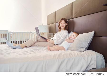 Baby and mother with a laptop working on a bed. 74879857