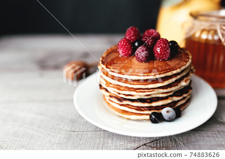 Juicy pancakes with berries and honey on a white plate, spoon, jar, wooden table 74883626
