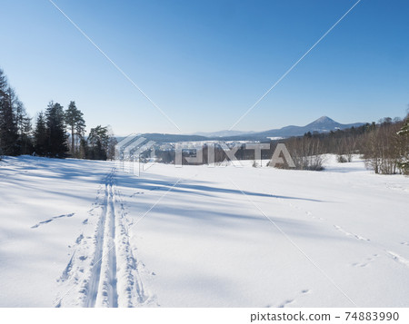 Winter landscape with view of village town Cvikov and ski run trail on snow-covered fields and snowy frozen forest and trees on sunny day, blue sky background 74883990