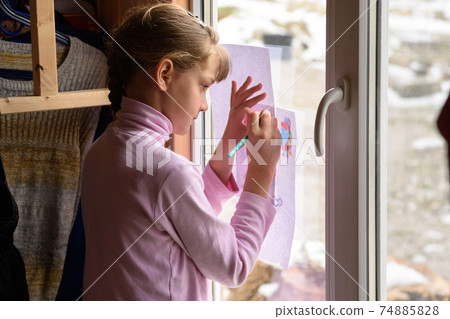 The girl draws a picture on a blank sheet by attaching the picture to the window glass 74885828