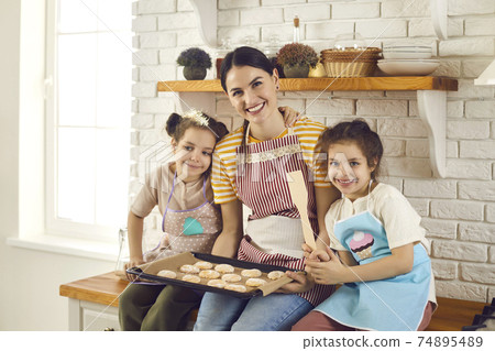 Portrait of happy young mom and little children in aprons with baking tray full of cookies Portrait of happy young mom and little children in aprons with baking tray full of cookies 74895489
