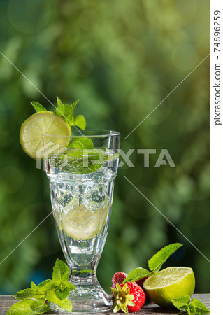 in a tall glass a refreshing drink with the addition of mint, lime and ice, against a background of greenery, next to strawberries and mint leaves in a tall glass a refreshing drink with the addition of mint, lime and ice, against a background of greenery, next to strawberries and mint leaves 74896259