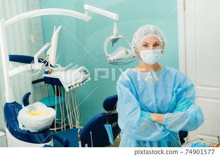 a female dentist wearing a medical mask and rubber gloves poses for the camera and folds her arms in her office a female dentist wearing a medical mask and rubber gloves poses for the camera and folds her arms in her office 74901577