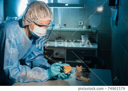 A masked and gloved dental technician works on a prosthetic tooth in his lab A masked and gloved dental technician works on a prosthetic tooth in his lab 74901579
