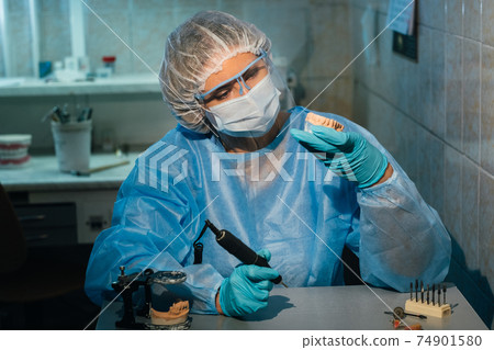 A masked and gloved dental technician works on a prosthetic tooth in his lab A masked and gloved dental technician works on a prosthetic tooth in his lab 74901580
