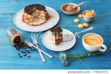 Round tiramisu, homemade no-bake traditional Italian sweet dessert. Blue table with cake, bowl with cocoa powder, round sponge biscuits, coffee beans and coffee with milk. Blue background. 74902971