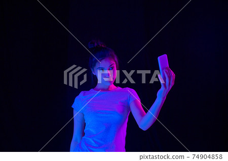 Young beautiful girl taking selfies isolated over dark background in neon light. 74904858