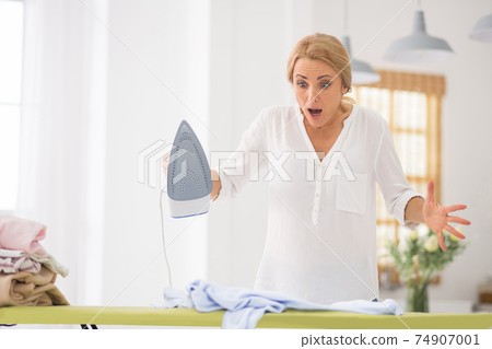 Pretty blonde woman standing near ironing table and looking shocked 74907001