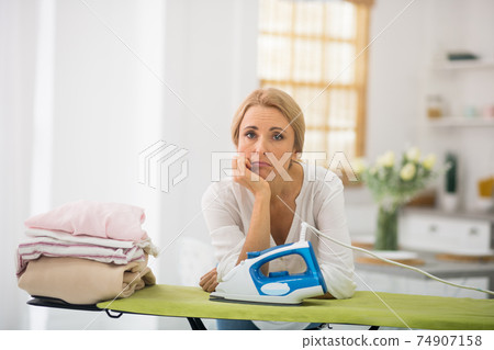 Blonde woman standing near the ironng table and looking sad 74907158
