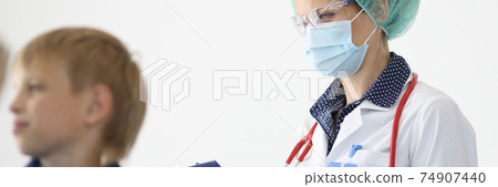 Doctor in protective medical mask writes readings on clipboard next to doctor listens with stethoscope to boy's breath. Doctor in protective medical mask writes readings on clipboard next to doctor listens with stethoscope to boy's breath. 74907440