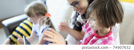 Woman is sitting on the couch, girl is sitting on her lap, they are looking at laptop next to children are playing. Woman is sitting on the couch, girl is sitting on her lap, they are looking at laptop next to children are playing. 74907632