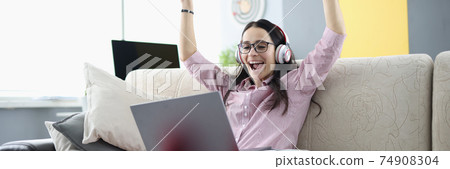Rejoicing woman is sitting on sofa with in headphones with laptop. Rejoicing woman is sitting on sofa with in headphones with laptop. 74908304