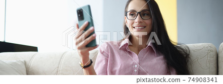 Woman with glasses sits on couch smiling and looks at smartphone screen 74908341