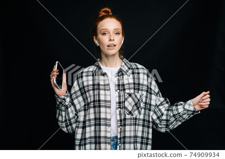 Cheerful red-haired young woman wearing wireless earphones holding phone and listening online music. 74909934