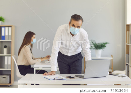 Adult man in a protective medical mask stands in the office against the background of his colleague. 74913664